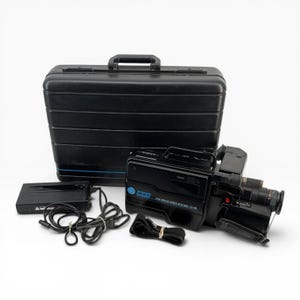 May include: A black GE VHS Movie Video System camcorder with a lens, a black hard-shell carrying case, a battery pack with a power cord, and a black strap. The camcorder features blue accents and text labels on the side, including the GE logo.