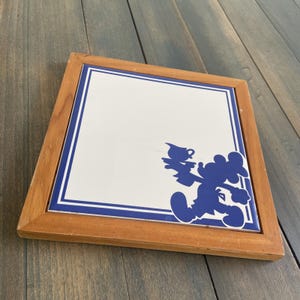 May include: Square wooden framed tile with a white center and blue border. A blue silhouette of Mickey Mouse holding a tea set is in the corner. The frame is made of light brown wood.