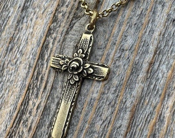 May include: A brass-toned metal cross pendant suspended from a matching link chain. The cross features an embossed floral design at the centre where the arms intersect, with textured, linear detailing along the length of each arm of the cross.