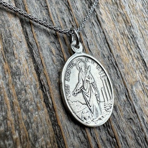 May include: Silver chain necklace with a round silver pendant featuring a depiction of Saint Agatha. The pendant has the inscription "S. AGATHA. O.R.P.N." around the edge.