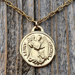 May include: Gold chain necklace with a round pendant featuring a detailed image of Saint Michael the Archangel. The pendant is inscribed with "SAINT" and "MICHAEL".