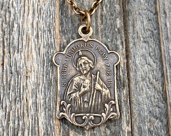 Op de afbeelding: Een bronzen metalen ketting met een rechthoekige religieuze hanger met versierde randen. De medaille toont een figuur die een staf en een klein hart vasthoudt, met de in reliëf aangebrachte tekst St. Jude Thaddeus Pray For Us.
