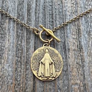 Op de afbeelding: Gouden ketting met een schuifgesp en een ronde hanger met een religieus beeld van de Maagd Maria met de tekst "O PUR MARIE CONCE SANS PECH PRIEZ NOUS QUI AVONS RECOURS A VOUS".
