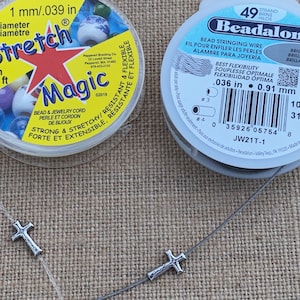 May include: Two spools of beading supplies are shown, one with clear Stretch Magic cord and the other with Beadalon bead stringing wire. Two silver cross charms are strung on the clear cord. The Stretch Magic spool is 1 mm diameter and 25 m long.