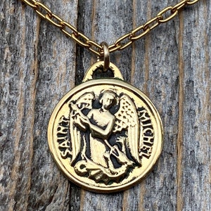 May include: Gold chain necklace with a round pendant featuring a depiction of Saint Michael the Archangel with a sword and a dragon. The pendant is inscribed with "Saint Michael" on the left side and "Michel" on the right side.