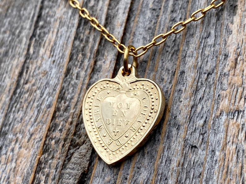 May include: Gold heart-shaped pendant necklace with a delicate chain. The pendant features an inscription that reads "Moy En Diev", surrounded by a decorative border.