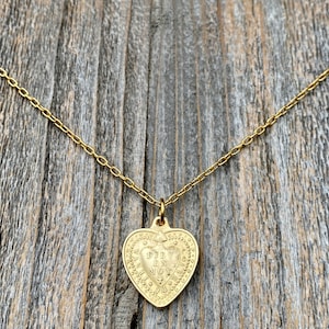 May include: Gold heart-shaped pendant necklace with the inscription "DIV IN NOY" on a gold chain.