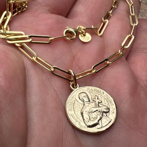 Può includere: Una collana a catena dorata con maglie a forma di graffetta e un ciondolo rotondo con l'immagine in rilievo di un santo che regge una croce. Il ciondolo reca la scritta "SAINT GERARD MAJELLA PRIEZ POUR NOUS" e include una piccola targhetta circolare vicino alla chiusura.