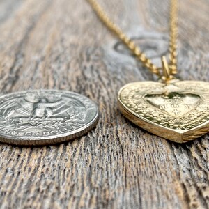 May include: A gold heart-shaped pendant necklace with a detailed design and the words "JESUS MY LOVE" inscribed inside. The pendant is lying on a wooden surface next to a US quarter.