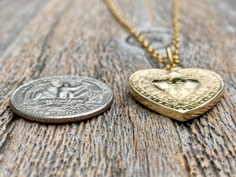 May include: A gold heart-shaped pendant necklace with a detailed design and the words "JESUS MY LOVE" inscribed inside. The pendant is lying on a wooden surface next to a US quarter.