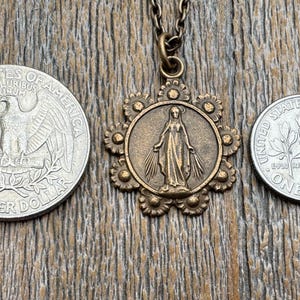 May include: A gold-toned pendant necklace featuring a detailed image of the Virgin Mary with a crown of stars. The pendant is surrounded by a decorative floral design with small beads.