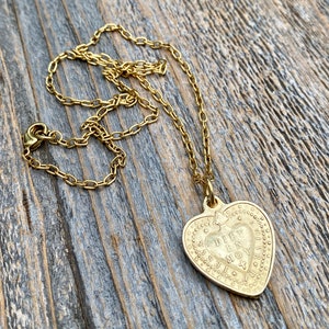 May include: A gold heart-shaped pendant necklace with a detailed inscription that reads "DIEU EST MON AMOUR". The necklace is on a gold chain and is lying on a wooden surface.