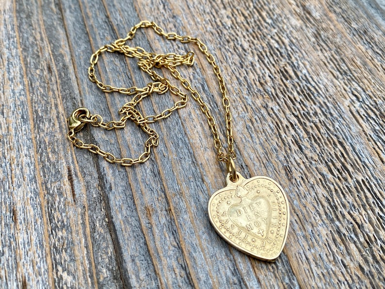 May include: A gold heart-shaped pendant necklace with a detailed inscription that reads "DIEU EST MON AMOUR". The necklace is on a gold chain and is lying on a wooden surface.