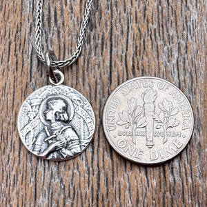 May include: A silver pendant necklace featuring a round medallion with a detailed relief of a saint, next to a US dime coin for size comparison. The pendant hangs from a braided chain. The background is a wooden surface.