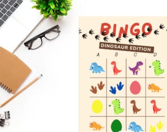 Könnte beinhalten: Eine "Dinosaurier Edition" Bingo-Spielkarte mit farbenfrohen Dinosaurier-Illustrationen und dem Wort "BINGO" oben. Die Karte liegt auf einer weißen Oberfläche mit einem Laptop, Notizbuch, Brille und einer kleinen Pflanze.