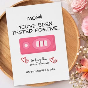 Może przedstawiać: Kartka na Dzień Matki z napisem "MOM!! YOU'VE BEEN TESTED POSITIVE..." i różową ilustracją testu ciążowego. Na kartce widnieje również napis "for being the coolest mom ever" i "HAPPY MOTHER'S DAY."