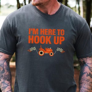 May include: Dark gray t-shirt with the text "I'M HERE TO HOOK UP" in orange. The shirt features an orange tractor graphic with checkered flags on either side. The tee shirt is a casual wear item.