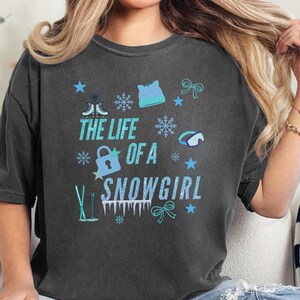 May include: Dark gray t-shirt with the text "THE LIFE OF A SNOWGIRL" in turquoise. The design includes snowflakes, stars, ice skates, a lock, a hat, a bow, and ski poles in turquoise and white.