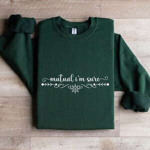 Puede incluir: Una sudadera de cuello redondo en color verde bosque oscuro con el texto blanco "mutual i'm sure" en una fuente cursiva, centrado sobre un elemento decorativo de l&iacute;nea con un peque&ntilde;o icono de copo de nieve en el centro.