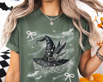 May include: Dark green t-shirt featuring a detailed illustration of a black witch hat adorned with pearls, feathers, and a buckle. The design includes white bats and bows, creating a Halloween theme. The shirt is a comfortable, casual style.