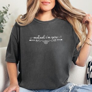 Puede incluir: Una camiseta gris carb&oacute;n de manga corta con el texto en blanco mutual i'm sure, centrado sobre un adorno decorativo y un peque&ntilde;o icono de copo de nieve. La camiseta tiene cuello redondo y un corte holgado con textura de tela lavada.