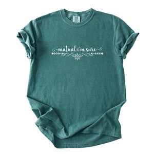 Puede incluir: Una camiseta de manga corta color verde azulado con el texto blanco "mutual i'm sure" centrado en el pecho. El texto est&aacute; flanqueado por adornos blancos decorativos y un peque&ntilde;o icono de copo de nieve, todo impreso en una fuente cursiva blanca.