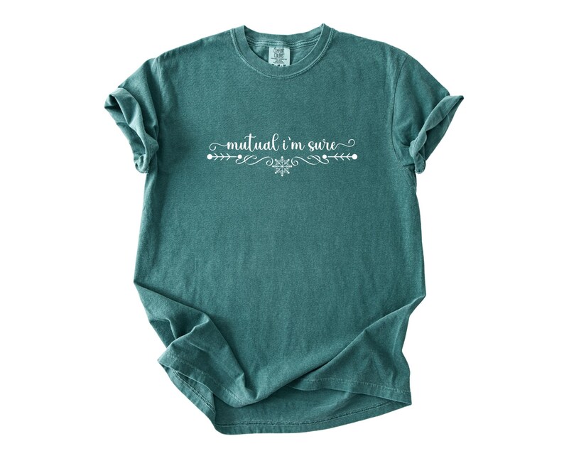 Puede incluir: Una camiseta de manga corta color verde azulado con el texto blanco "mutual i'm sure" centrado en el pecho. El texto est&aacute; flanqueado por adornos blancos decorativos y un peque&ntilde;o icono de copo de nieve, todo impreso en una fuente cursiva blanca.