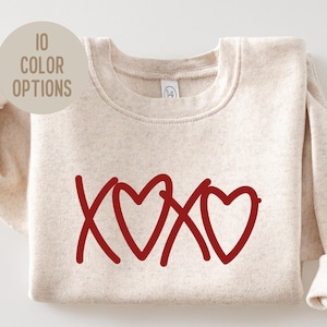 May include: A beige sweatshirt with the red text "XOXO" and three hearts. The image includes a circular graphic that says "10 COLOR OPTIONS". The sweatshirt is folded with the sleeves visible.