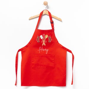 May include: Red kitchen apron with a pocket, featuring a design of wooden cooking utensils and green leaves. The name "Henry" is embroidered in white on the apron.