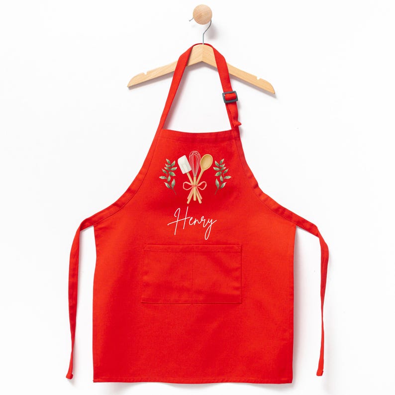 May include: Red kitchen apron with a pocket, featuring a design of wooden cooking utensils and green leaves. The name "Henry" is embroidered in white on the apron.