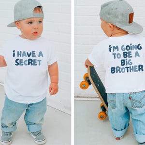 May include: A toddler wearing a white t-shirt with the text "I HAVE A SECRET" in blue lettering. The child is also wearing light blue jeans, gray sneakers, and a gray baseball cap. The back of the shirt says "I'M GOING TO BE A BIG BROTHER".