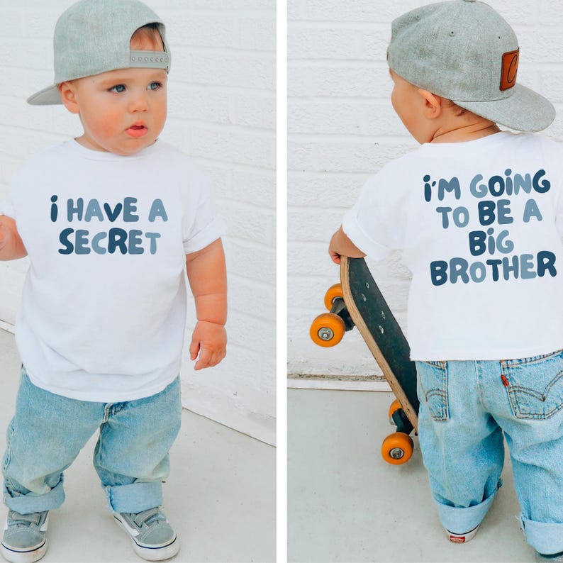 May include: A toddler wearing a white t-shirt with the text "I HAVE A SECRET" in blue lettering. The child is also wearing light blue jeans, gray sneakers, and a gray baseball cap. The back of the shirt says "I'M GOING TO BE A BIG BROTHER".