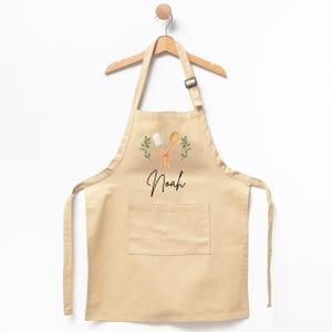 May include: A beige cotton apron with a pocket, featuring a graphic of crossed wooden spoons and green sprigs, with the name "Noah" printed below.