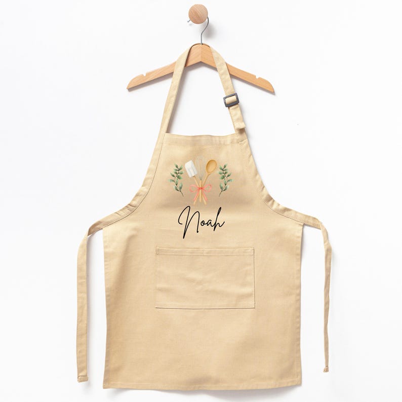May include: A beige cotton apron with a pocket, featuring a graphic of crossed wooden spoons and green sprigs, with the name "Noah" printed below.