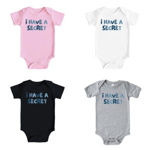 May include: Four baby onesies in pink, white, black, and gray. Each onesie has the text "I HAVE A SECRET" in blue lettering. The onesies are short-sleeved and have a snap closure at the bottom.