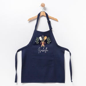 May include: A navy blue apron with a pocket, featuring a white embroidered design of crossed kitchen utensils, a red ribbon, and green leaves. The name "Isabella" is embroidered below the design.