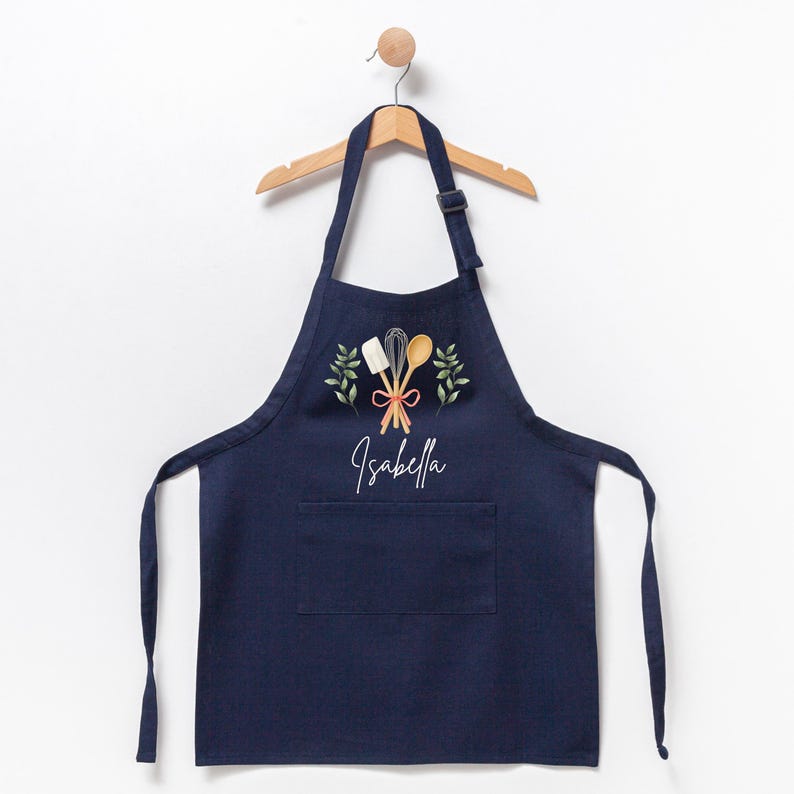 May include: A navy blue apron with a pocket, featuring a white embroidered design of crossed kitchen utensils, a red ribbon, and green leaves. The name "Isabella" is embroidered below the design.