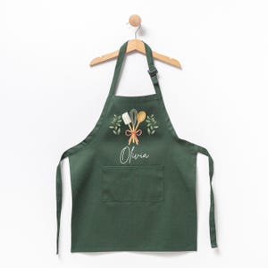 May include: A dark green apron with a pocket and adjustable straps. The apron has a design of kitchen utensils, greenery, and the name "Olivia" printed on it.