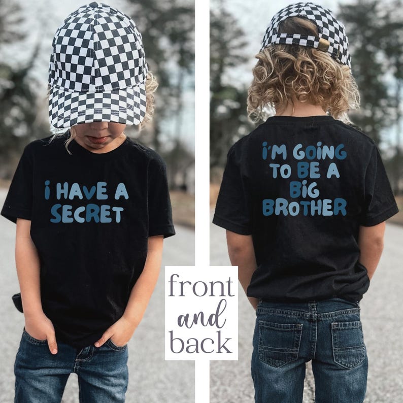 May include: A black t-shirt with the text "I HAVE A SECRET" in blue on the front and "I'M GOING TO BE A BIG BROTHER" on the back. The child is wearing a black and white checkered baseball cap and blue jeans.