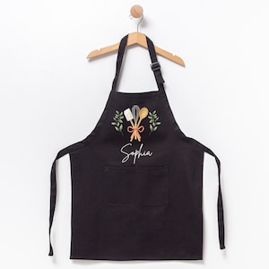 May include: Black apron with a pocket, featuring a design of kitchen utensils and green leaves. The name "Sophia" is embroidered in white on the apron.