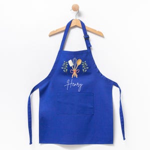 May include: A blue kitchen apron with a pocket, featuring a white design of kitchen utensils and greenery, with the name "Henry" written in white script.