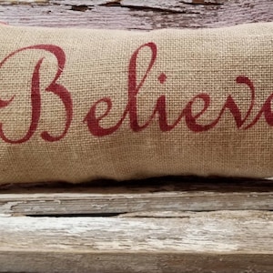 May include: A burlap pillow with the word "Believe" printed in red cursive lettering. The pillow is sitting on a rustic wooden bench with green and red Christmas ornaments and pine branches.