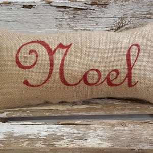 May include: A burlap pillow with the word "Noel" written in red cursive lettering.