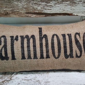 Pu&ograve; includere: Un cuscino di juta marrone con la parola "Farmhouse" stampata in nero.