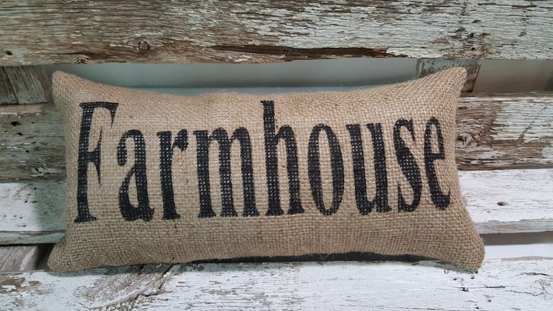Pu&ograve; includere: Un cuscino di juta marrone con la parola "Farmhouse" stampata in nero.