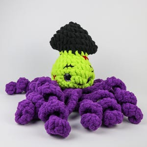 Könnte beinhalten: Ein handgefertigtes, plüschiges Frankenstein-Monster-Spielzeug mit einem limettengrünen Kopf, schwarzen Haaren und lila Tentakeln. Das Monster hat gestickte Gesichtszüge und eine kleine rote Naht auf dem Kopf. Das Spielzeug ist aus weichem, dickem Garn gefertigt.