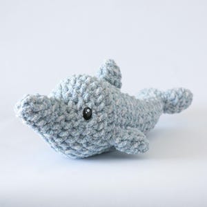 May include: A light blue crocheted dolphin toy with a textured finish. The dolphin has a rounded body, a small dorsal fin, and flippers. It features a black button eye on each side of its head. The toy is designed for children.