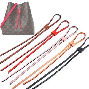 Pode incluir: Uma bolsa marrom com alça e cordão rosa, junto com várias substituições de cordão de couro em várias cores: marrom, vermelho, rosa, bege e preto. A bolsa e as alças estão em um fundo branco.