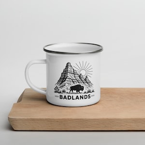 May include: A white enamel mug with a silver rim and a black graphic print. The design features a stylized mountain landscape, a sun, a bison silhouette, and the text BADLANDS centered below the illustration in a clean, bold sans-serif font.