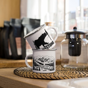 May include: Two white enamel mugs with black designs. The top mug features a black bear silhouette with the word "Stone" printed on the side. The bottom mug features a black mountain range silhouette with a river and trees.